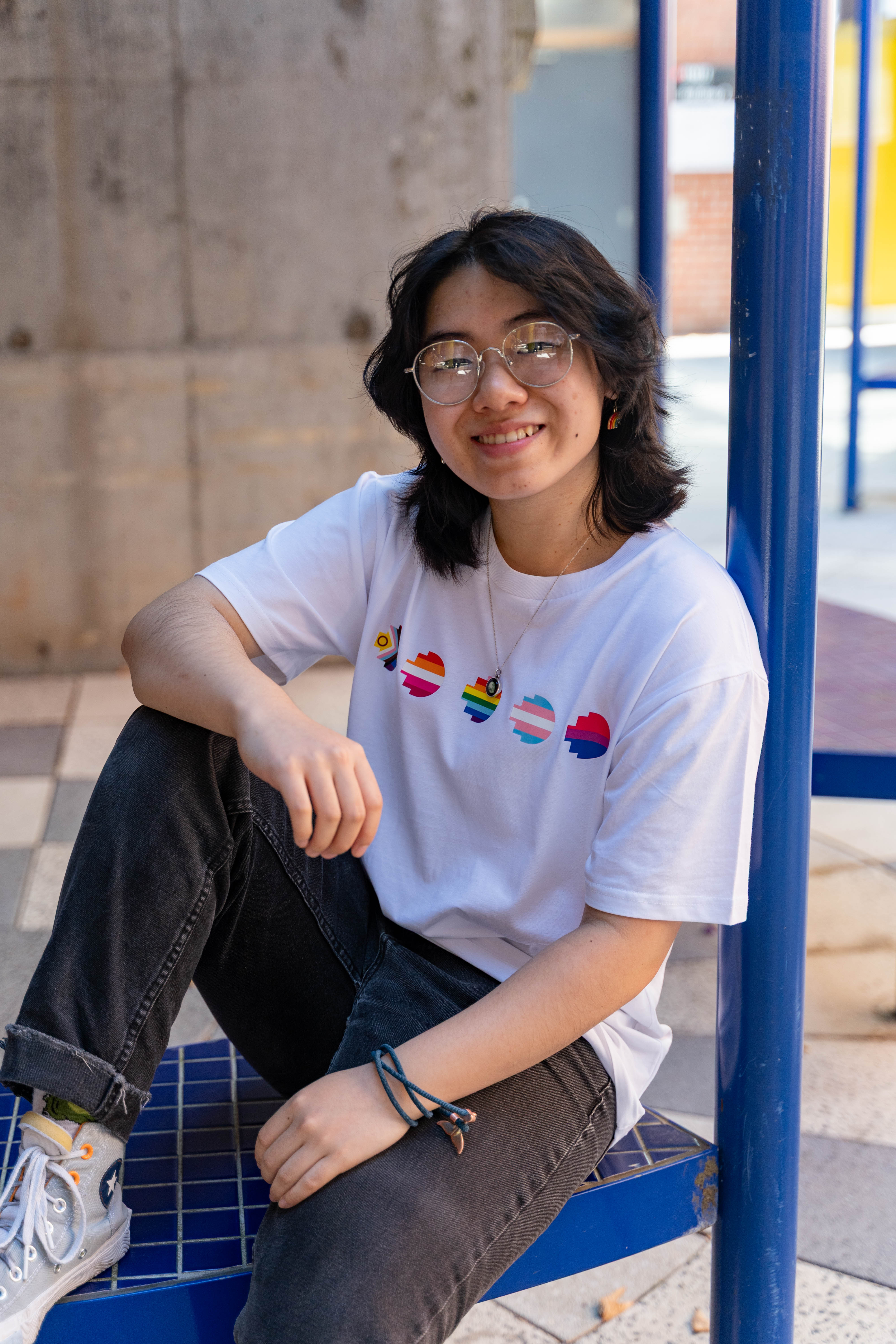 Model wears the Pride Unity Tee in limited edition white, featuring colourful pride emblem prints on the chest, displaying the plain back design against a brick wall background. Available at the RMIT Store.